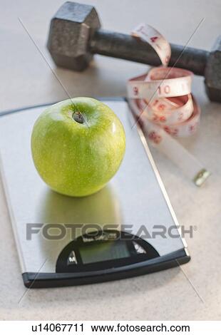 Stock Image - Green apple on weight scale, tape measure and exercise weight in background. Fotosearch