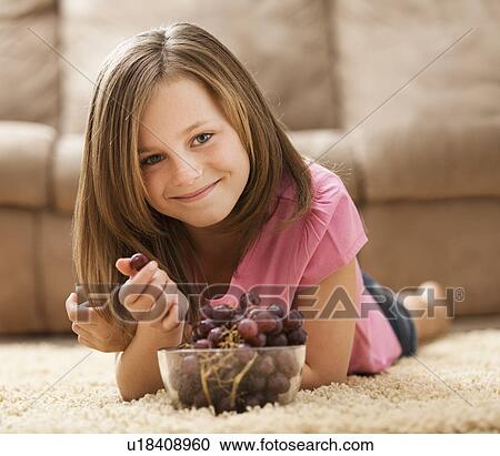 Stock Image - Portrait of girl (10-11) lying on rug, eating red grapes. Fotosearch