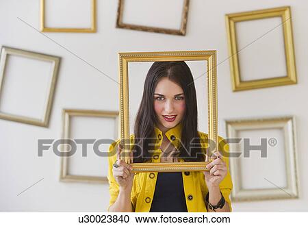 Stock Image - Portrait of woman holding picture frame over face. Fotosearch