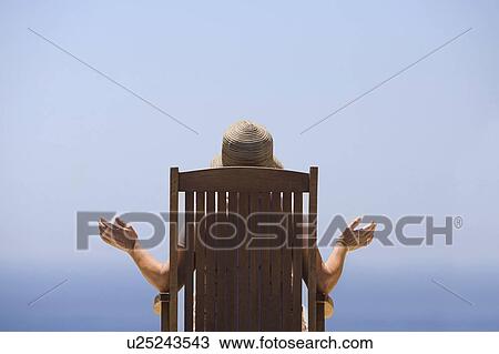 Stock Image - Rear view of woman sitting on wooden chair, facing sea. Fotosearch