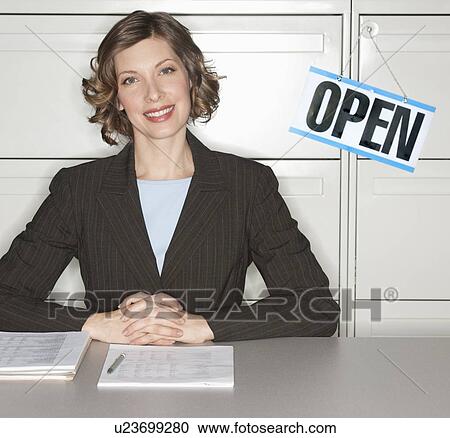 Stock Image - Smiling businesswoman sitting at desk next to open sign. Fotosearch
