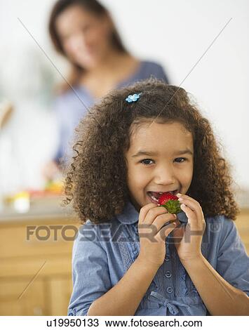 Smiling girl (6-7) eating strawberries with defocused woman in background View Large Photo Image Stock Image - Smiling girl (6-7) eating strawberries with defocused woman in background. Fotosearch
