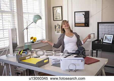 Stock Photography - Woman sitting in office room, holding cup of tea, with arms stretched. Fotosearch
