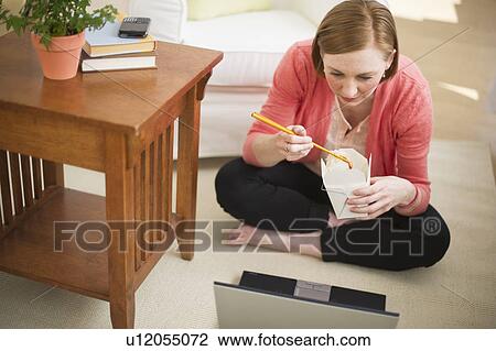 Stock Image - Woman sitting on floor, using laptop and eating take out food. Fotosearch
