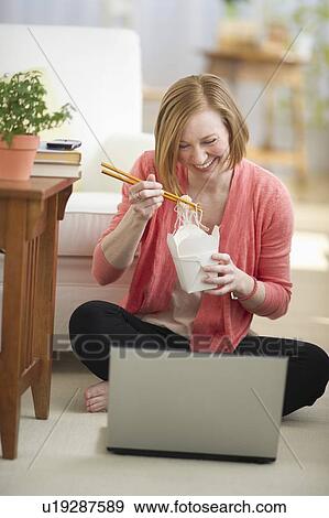Stock Photo - Woman sitting on floor, using laptop and eating take out food. Fotosearch