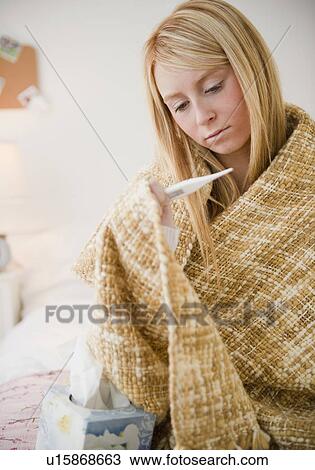 Stock Image - Woman wrapped in blanket measuring temperature. Fotosearch