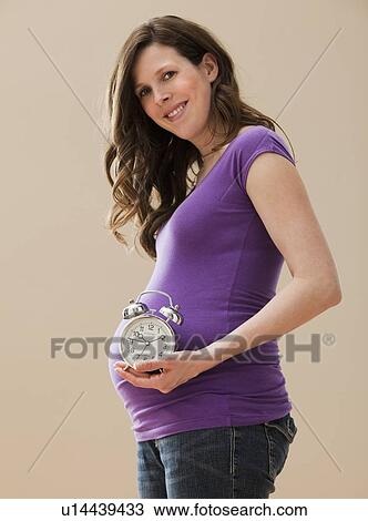 Stock Image - Young pregnant woman holding alarm clock next to belly. Fotosearch