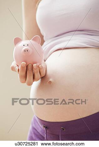 Stock Image - Young pregnant woman holding pink piggybank. Fotosearch
