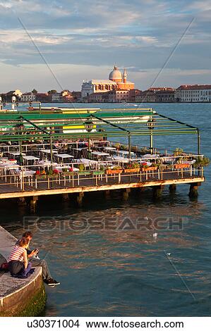 Aerial view of Canale della Giudecca, looking towards church of Il Redentore, in late afternoon golden sunlight, with empty outdoor restaurant, and couple sitting on quayside in foreground, looking at their photos on camera, Venice, Northern Italy View Large Photo Image Picture - Aerial view of Canale della Giudecca, looking towards church of Il Redentore, in late afternoon golden sunlight, with empty outdoor restaurant, and couple sitting on quayside in foreground, looking at their photos on camera, Venice, Northern Italy. Fotosearch
