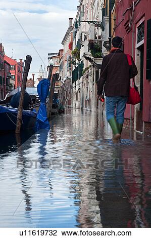 Stock Image - Aqua Alta: back view of man wearing green wellington boots wading through water along canal, boats moored alongside canal with reflections of buildings in water, and bell tower in distance, sunny autumn morning in Venice, Northern Italy. Fotosearch