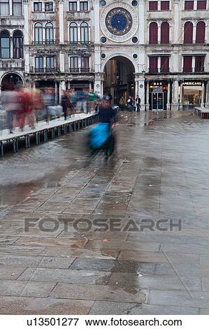 Stock Photo - Aqua Alta flooding in Piazza San Marco: motion blur of pedestrians walking on duckboards, man wheeling blue-covered wheelbarrow, and Clock Tower and loggia of the Procuratie behind, Venice, Northern Italy. Fotosearch
