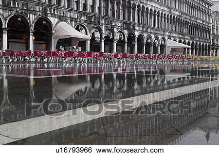 Stock Photograph - Aqua Alta flooding in Piazza San Marco: desaturated image highlighting empty red and yellow chairs, and grey facade and loggia of the Procuratie behind, reflected in the water, Venice, Northern Italy. Fotosearch