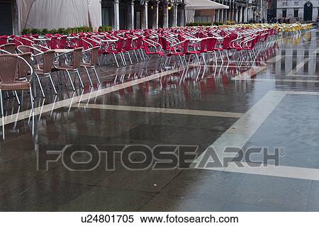 Stock Photography - Aqua Alta flooding in Piazza San Marco: empty red and yellow chairs, and grey facade and loggia of the Procuratie behind, reflected in the water, Venice, Northern Italy. Fotosearch