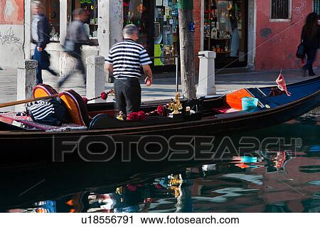 Stock Image - Back view of gondolier in moored gondola, cleaning his gondola, with motion blur of passers by, reflections in water, sunny autumn morning in Venice, Northern Italy. Fotosearch