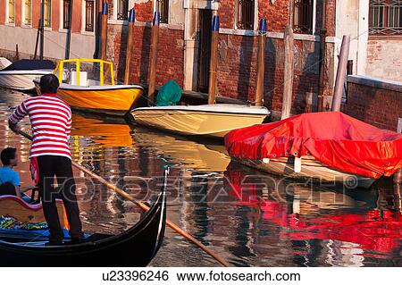 Stock Photograph - Back view of gondolier in red and white striped shirt, rowing past moored boats in late afternoon golden light, reflections in water, autumn in Venice, Northern Italy. Fotosearch