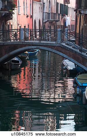 Boats moored alongside canal under stone bridge with iron railings, reflections of palace buildings in water, sunny autumn morning in Venice, Northern Italy View Large Photo Image Stock Photography - Boats moored alongside canal under stone bridge with iron railings, reflections of palace buildings in water, sunny autumn morning in Venice, Northern Italy. Fotosearch