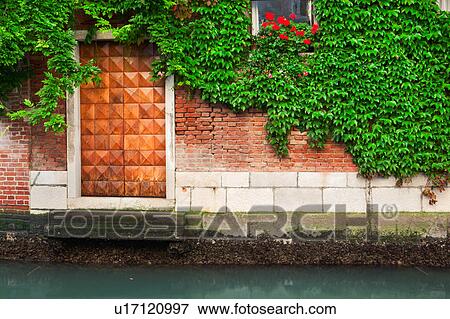 Stock Photo - Canal-side doorway to home, with wooden door, window with red geraniums, and ivy trailing across red brick wall, Venice, Northern Italy. Fotosearch