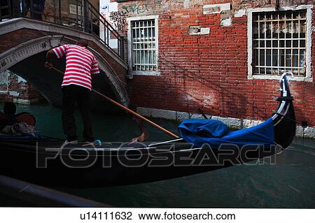 Stock Image - Close up: back view of gondolier in red and white striped shirt bending to pass gondola under arch of red brick bridge, sunny autumn morning in Venice, Northern Italy. Fotosearch