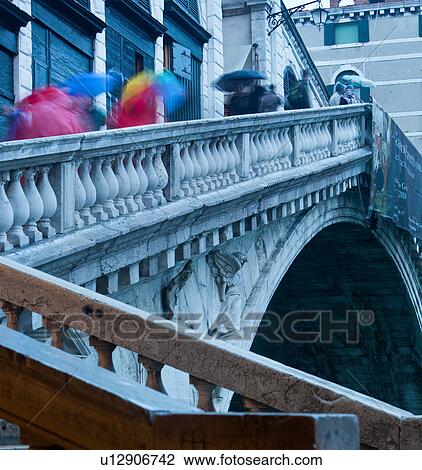 Stock Image - Close up of Rialto Bridge from below, in rain at twilight, with motion blur of pedestrians on outer stairway, with bright red coats and multicoloured umbrellas, against white stone balustrade, Venice, Northern Italy. Fotosearch