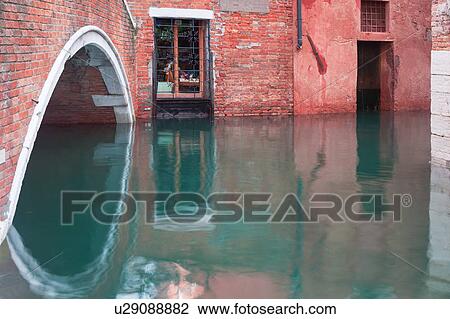 Stock Image - Close up view of red brick arch of bridge and wall with shop window and door, canal water flooding and reflection, at Aqua Alta, Venice, Northern Italy. Fotosearch