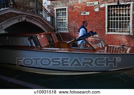 Stock Photo - Close up: white and mahogany motorboat water taxi, and taxi driver steering under arch of red brick bridge, sunny autumn morning in Venice, Northern Italy. Fotosearch