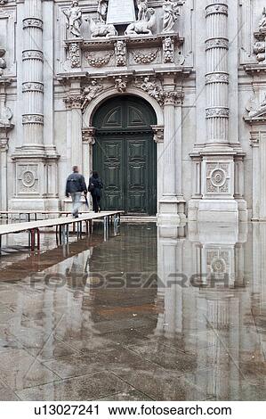 Stock Image - Detail of ornate Baroque facade and doorway of San Moise church with pedestrian couple walking single file on duckboards, all reflected in flood waters of Aqua Alta, Venice, Northern Italy. Fotosearch