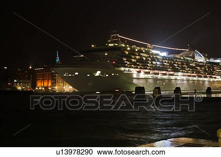 Stock Image - Huge cruise liner departing at night through the Canale della Giudecca, with Stucky Mill and buildings of Giudecca dwarfed behind, night illuminations, Venice Northern Italy. Fotosearch
