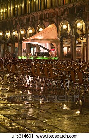 Piazza San Marco at night, with Aqua Alta flooding, lights of loggia reflected in puddles, rows of empty red and yellow chairs, empty orchestra, with grand piano, and motion blur of few passersby, Venice, Northern Italy View Large Photo Image Stock Image - Piazza San Marco at night, with Aqua Alta flooding, lights of loggia reflected in puddles, rows of empty red and yellow chairs, empty orchestra, with grand piano, and motion blur of few passersby, Venice, Northern Italy. Fotosearch