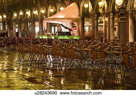 Stock Image - Piazza San Marco at night, with Aqua Alta flooding, lights of loggia reflected in puddles, rows of empty red and yellow chairs, empty orchestra, with grand piano, and motion blur of few passersby, Venice, Northern Italy. Fotosearch