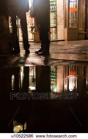 Piazza San Marco at night, with Aqua Alta flooding, detail of reflection of legs of two pedestrians standing, and lights of loggia souvenir shop reflected in puddles, Venice, Northern Italy View Large Photo Image Stock Photograph - Piazza San Marco at night, with Aqua Alta flooding, detail of reflection of legs of two pedestrians standing, and lights of loggia souvenir shop reflected in puddles, Venice, Northern Italy. Fotosearch