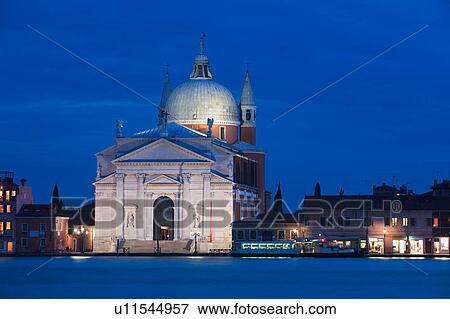 Stock Photo - View of church of Il Redentore, at twilight, with motion blur of passenger ferry, and water of Canale della Giudecca, lights illuminated along quayside, autumn in Venice, Northern Italy. Fotosearch