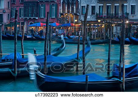 View of Grand Canal at twilight, motion blur of moored gondolas in foreground, gondoliers working on opposite side in front of illuminated shops and restaurants, autumn evening in Venice, Northern Italy View Large Photo Image Stock Image - View of Grand Canal at twilight, motion blur of moored gondolas in foreground, gondoliers working on opposite side in front of illuminated shops and restaurants, autumn evening in Venice, Northern Italy. Fotosearch