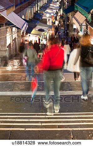 Stock Photo - View of Rialto Bridge central street from above, in rain at twilight, with motion blur of pedestrians on striped stairway, with bright red coat and multicoloured umbrellas, illuminated shops, Venice, Northern Italy. Fotosearch