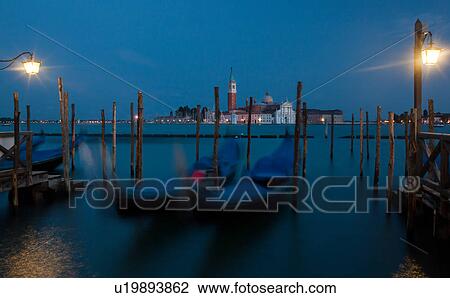 Stock Image - View of San Marco basin, looking past moored gondolas with motion blur in foreground, to San Giorgio Maggiore church and nearby harbour, dusk with lighthouse shining, lights of Giudecca, autumn in Venice, Northern Italy. Fotosearch