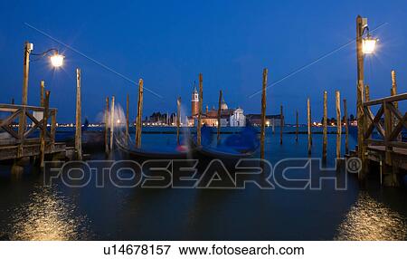 Stock Photo - View of San Marco basin, looking past moored gondolas with motion blur in foreground, to San Giorgio Maggiore church and nearby harbour, dusk with lighthouse shining, lights of Giudecca, autumn in Venice, Northern Italy. Fotosearch