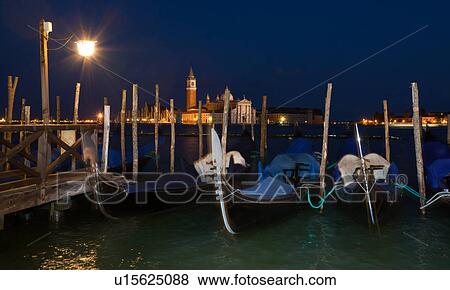 Stock Photo - View of San Marco basin, looking past moored gondolas with motion blur in foreground, to San Giorgio Maggiore church and nearby harbour, dusk with lighthouse shining, lights of Giudecca, autumn in Venice, Northern Italy. Fotosearch