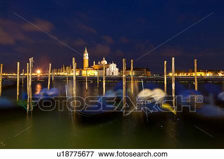 Stock Photo - View of San Marco basin, looking past moored gondolas with motion blur in foreground, to San Giorgio Maggiore church and nearby harbour, dusk with lighthouse shining, lights of Giudecca, autumn in Venice, Northern Italy. Fotosearch
