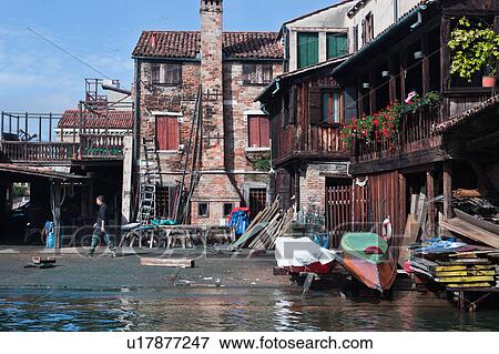 Stock Photo - View of San Trovaso gondola workshop with man striding across courtyard to shed, upturned gondolas in front of red brick building, sunny autumn morning in Venice, Northern Italy. Fotosearch