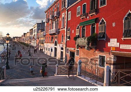Stock Image - View of Zattere, walkway along Canale della Giudecca at sunset, with motion blur of pedestrians and lovely architecture of palazzo buildings in golden light, autumn in Venice, Northern Italy. Fotosearch