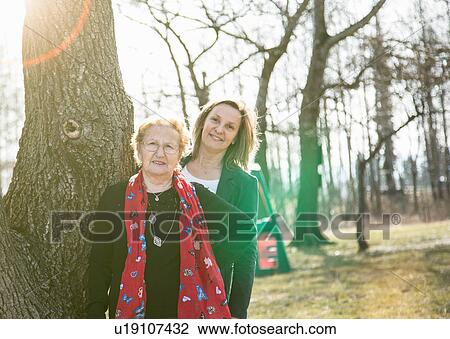 Adult daughter standing behind mother by tree looking at camera smiling View Large Photo Image Stock Image - Adult daughter standing behind mother by tree looking at camera smiling. Fotosearch