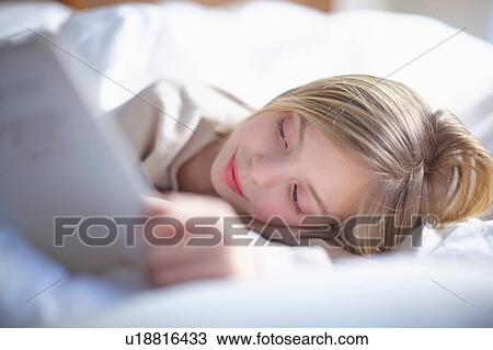 Stock Image - Blond haired girl lying in bed reading a book. Fotosearch