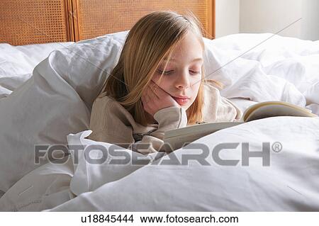 Picture - Blond haired girl reading book in bed. Fotosearch