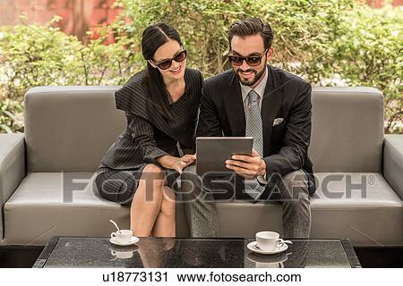 Stock Image - Businessman and woman reading digital tablet on hotel garden sofa, Dubai, United Arab Emirates. Fotosearch