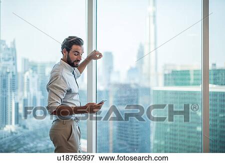 Stock Photo - Businessman reading smartphone text at window with view of Burj Khalifa, Dubai, United Arab Emirates. Fotosearch