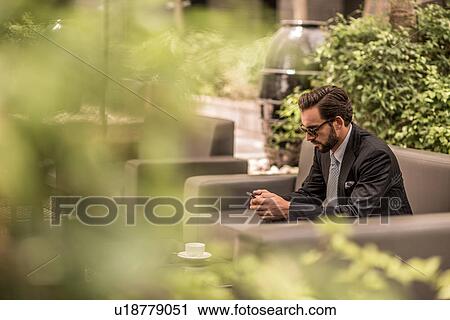 Stock Image - Businessman reading smartphone texts on hotel garden sofa, Dubai, United Arab Emirates. Fotosearch