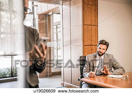 Stock Photo - Businessman sitting at desk talking to business woman holding smartphone. Fotosearch