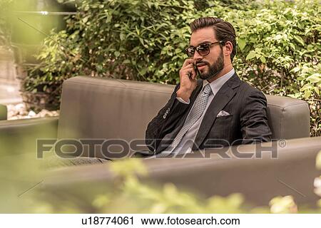 Stock Image - Businessman talking on smartphone on hotel garden sofa, Dubai, United Arab Emirates. Fotosearch