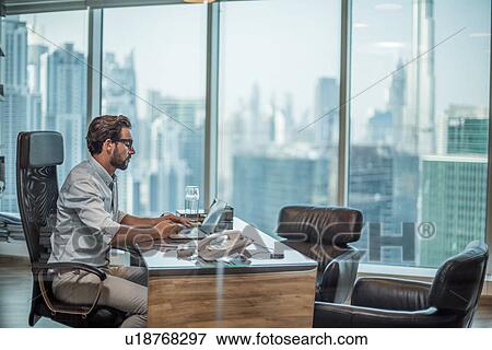 Stock Photo - Businessman using laptop at desk with window view of Burj Khalifa, Dubai, United Arab Emirates. Fotosearch