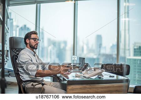 Stock Photograph - Businessman using smartphone at desk with window view of Burj Khalifa, Dubai, United Arab Emirates. Fotosearch