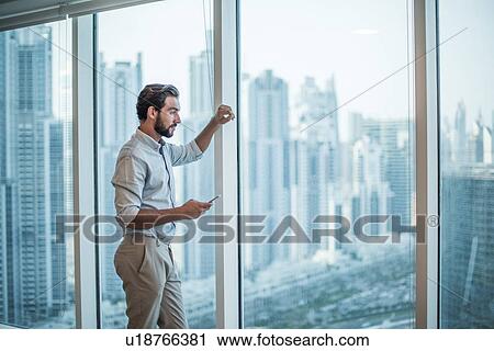 Stock Image - Businessman with smartphone staring through window with skyscraper view, Dubai, United Arab Emirates. Fotosearch
